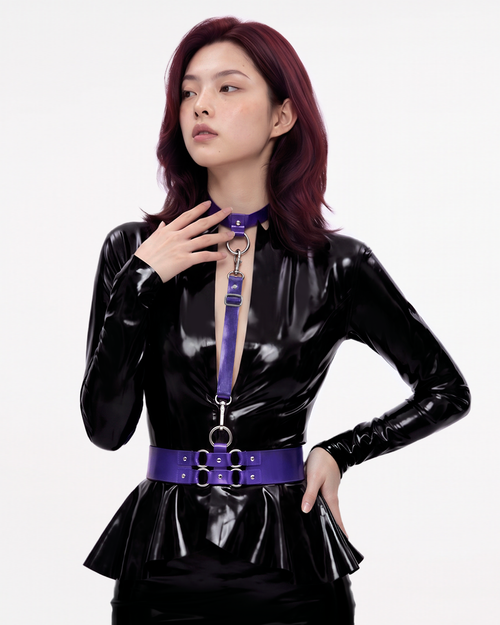 A model shown from the front wearing The Aurora Harness Set in metallic purple latex over a black latex dress, featuring a buckled choker, vertical chest strap, and a structured waist belt, accented with silver metallic O-rings and hardware.