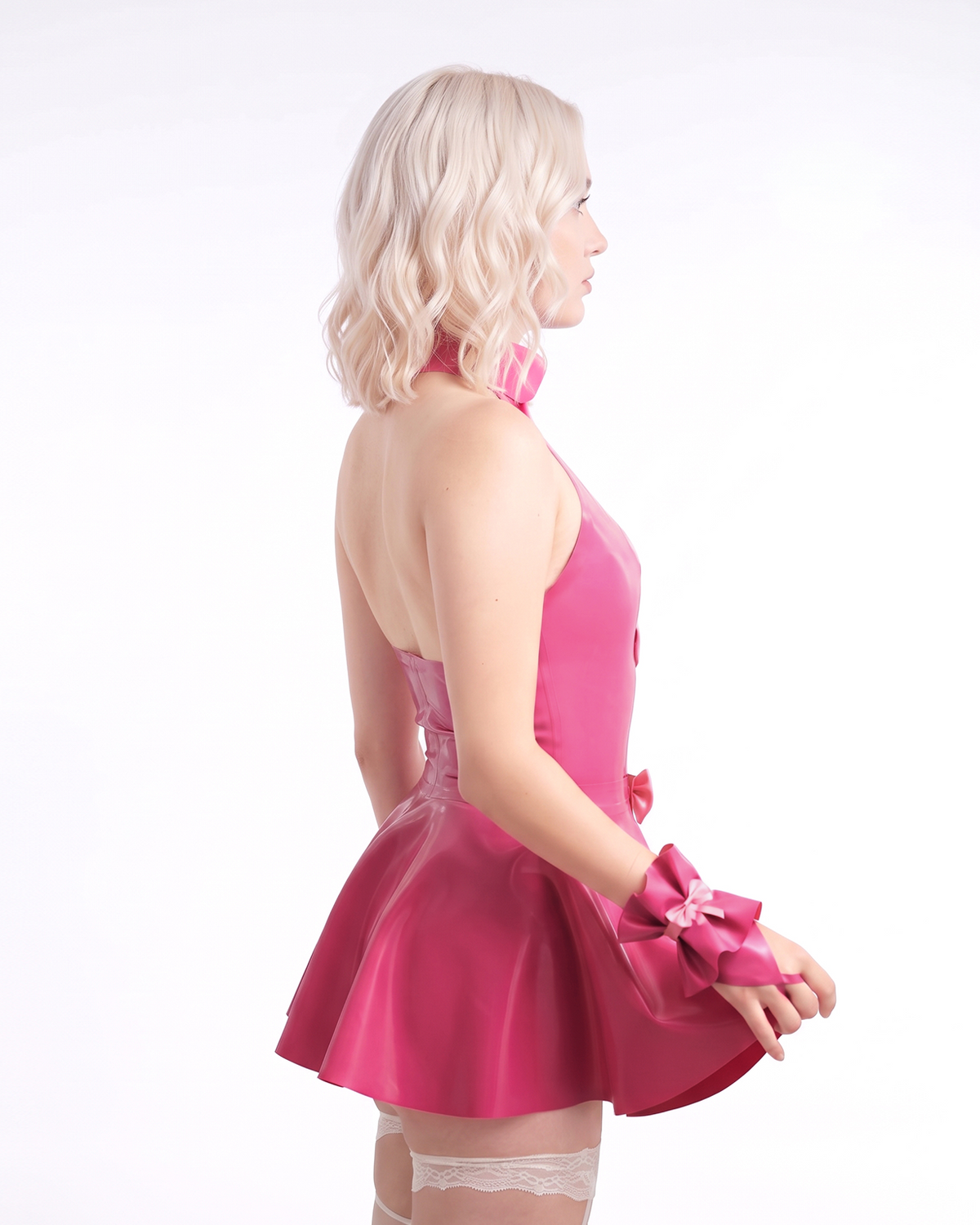 A model shown from the side wearing a high-shine vivid fuchsia latex multi-piece ensemble by Ribcage, featuring a collared halter bodysuit with a circular cutout and vertical bows, a separate flared peplum skirt overlay, and matching The Eloise Hand Cuffs, styled with white stockings.