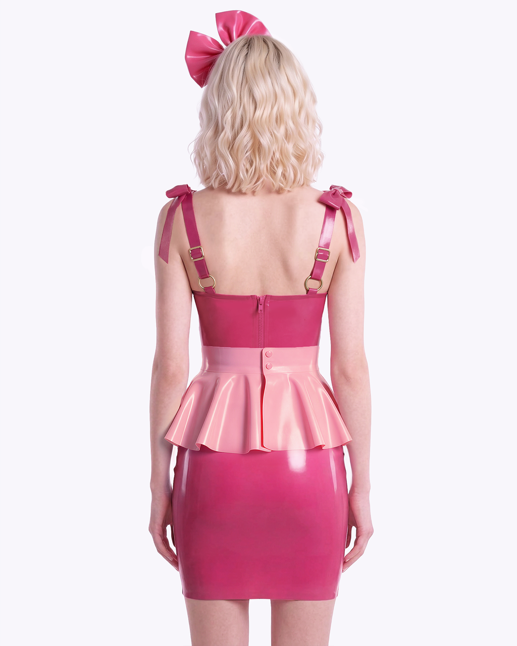 A model shown from the back wearing a high-shine color-blocked latex three-piece suit by Ribcage, highlighting the vivid fuchsia slip dress with a back zipper closure, the metallic pink peplum overlay, and the vivid fuchsia oversized bow headband.