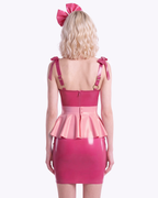 A model shown from the back wearing a high-shine color-blocked latex three-piece suit by Ribcage, highlighting the vivid fuchsia slip dress with a back zipper closure, the metallic pink peplum overlay, and the vivid fuchsia oversized bow headband.