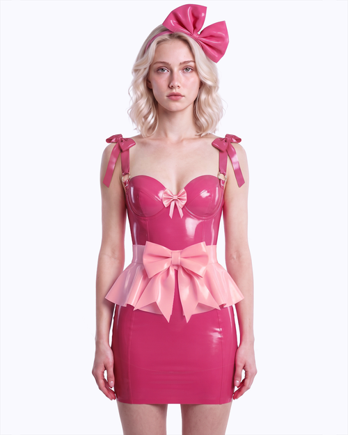 A model shown from the front wearing a high-shine color-blocked latex three-piece suit by Ribcage, featuring a vivid fuchsia slip dress with a metallic pink center bow, a contrasting metallic pink peplum overlay with a prominent front bow, and a vivid fuchsia oversized bow headband.