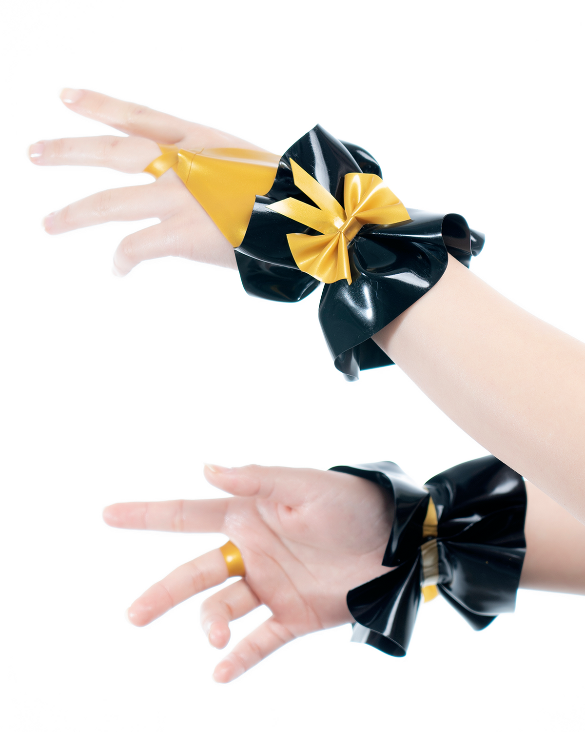 A model shown wearing a pair of high-shine metallic gold and black latex hand cuffs by Ribcage, featuring a flared ruffled cuff design, a contrasting black bow, and an integrated finger loop.