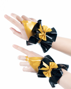 A model shown wearing a pair of high-shine metallic gold and black latex hand cuffs by Ribcage, featuring a flared ruffled cuff design, a contrasting black bow, and an integrated finger loop.