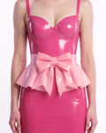 A model shown from the front wearing a high-shine metallic pink latex peplum overlay by Ribcage, featuring a dramatic oversized structural bow at the waist and a flared, softly pleated drape, styled over a black latex corset ensemble.