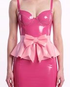A model shown from the front wearing a high-shine metallic pink latex peplum overlay by Ribcage, featuring a dramatic oversized structural bow at the waist and a flared, softly pleated drape, styled over a black latex corset ensemble.