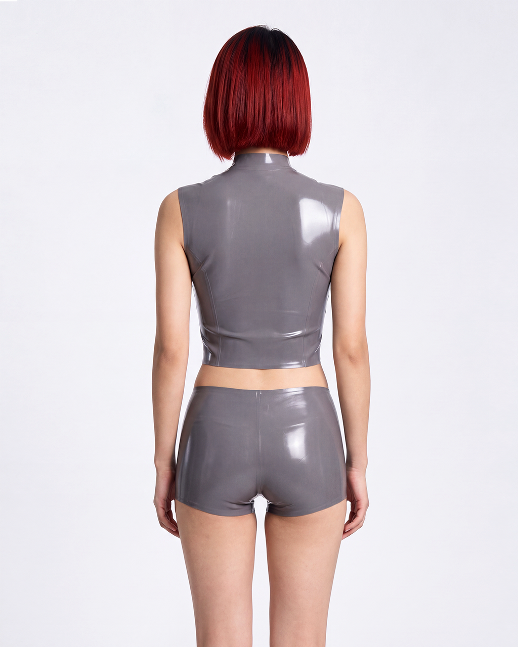 A model shown from the back wearing a high-shine metallic taupe-grey latex two-piece ensemble by Ribcage, featuring a mock-neck sleeveless crop top and matching high-waisted shorts in a compressive second-skin fit.