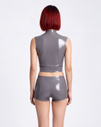 A model shown from the back wearing a high-shine metallic taupe-grey latex two-piece ensemble by Ribcage, featuring a mock-neck sleeveless crop top and matching high-waisted shorts in a compressive second-skin fit.
