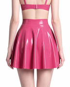 A model shown from the back wearing a high-shine vivid fuchsia latex short skirt by Ribcage, highlighting the seamless flared A-line silhouette and voluminous drape, styled with a matching crop top.