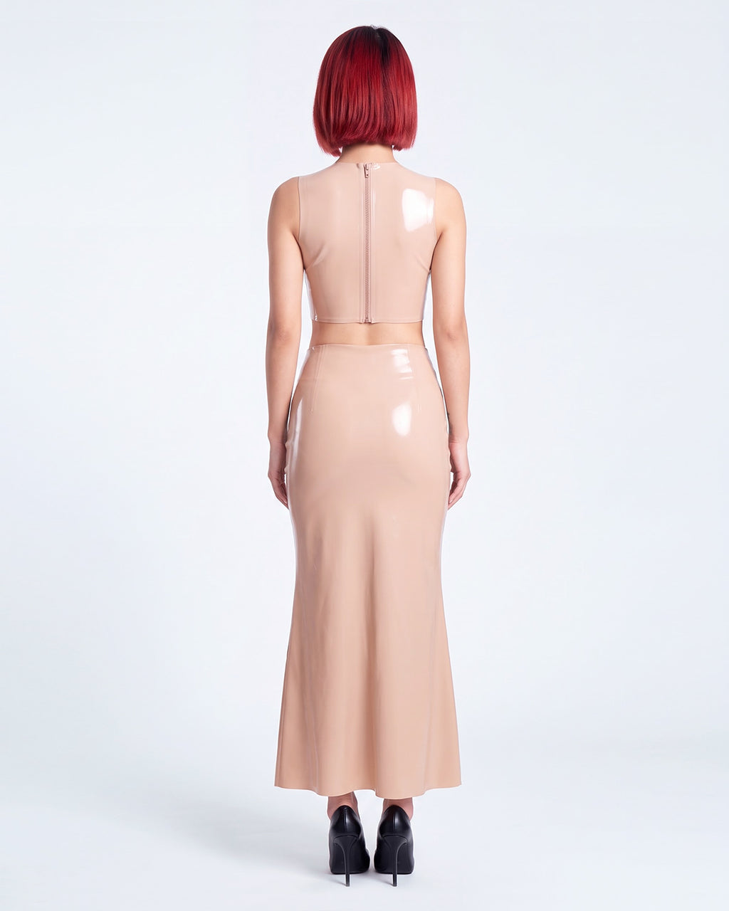 A model shown from the back wearing a high-shine nude latex two-piece ensemble by Ribcage, featuring a minimalist sleeveless crop top, a high-waisted maxi skirt, and a compressive second-skin fit.