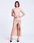 A model shown from the front wearing a high-shine nude latex two-piece ensemble by Ribcage, featuring a minimalist sleeveless crop top, a high-waisted maxi skirt with a dramatic side slit and a polished metal buckle accent, and a compressive second-skin fit.