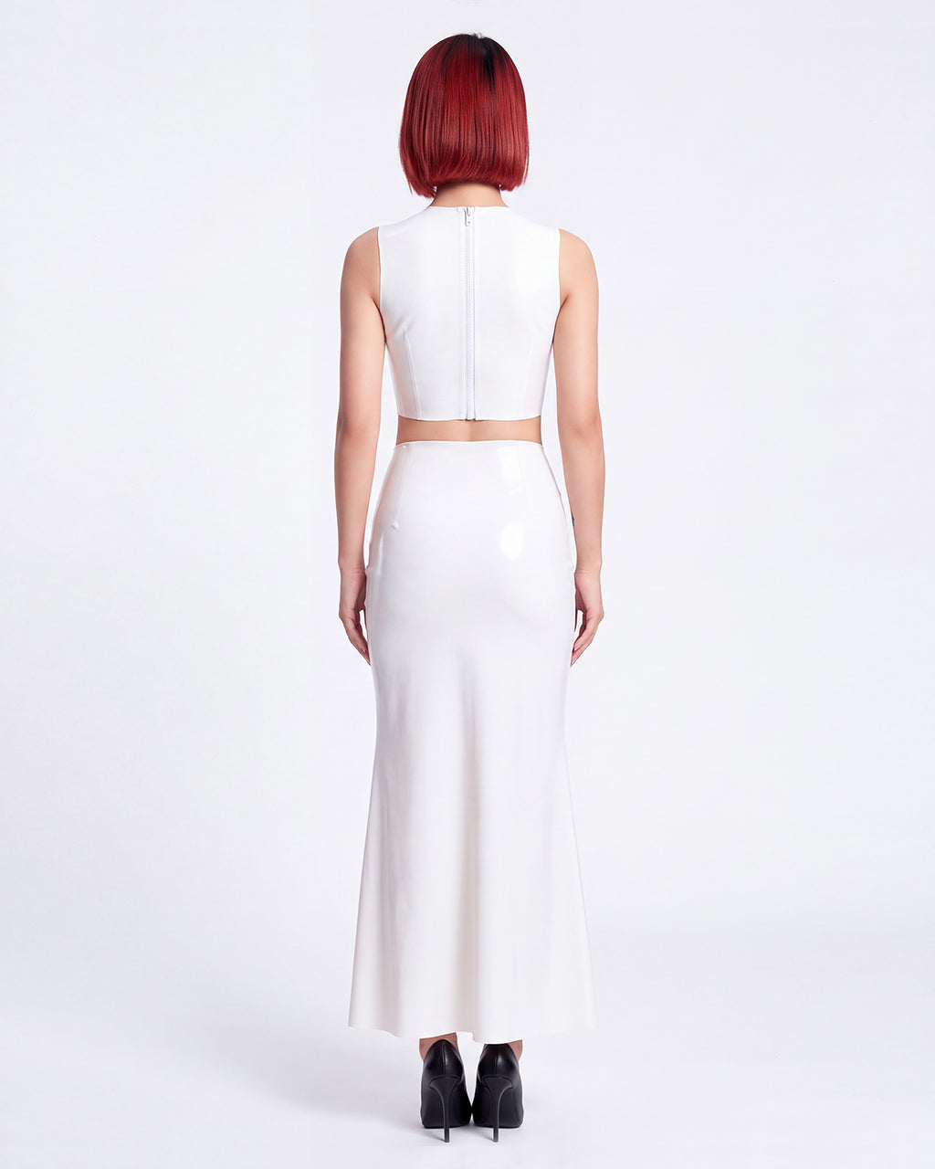 A model shown from the back wearing a high-shine white latex two-piece ensemble by Ribcage, featuring a minimalist sleeveless crop top, a high-waisted maxi skirt, and a compressive second-skin fit.