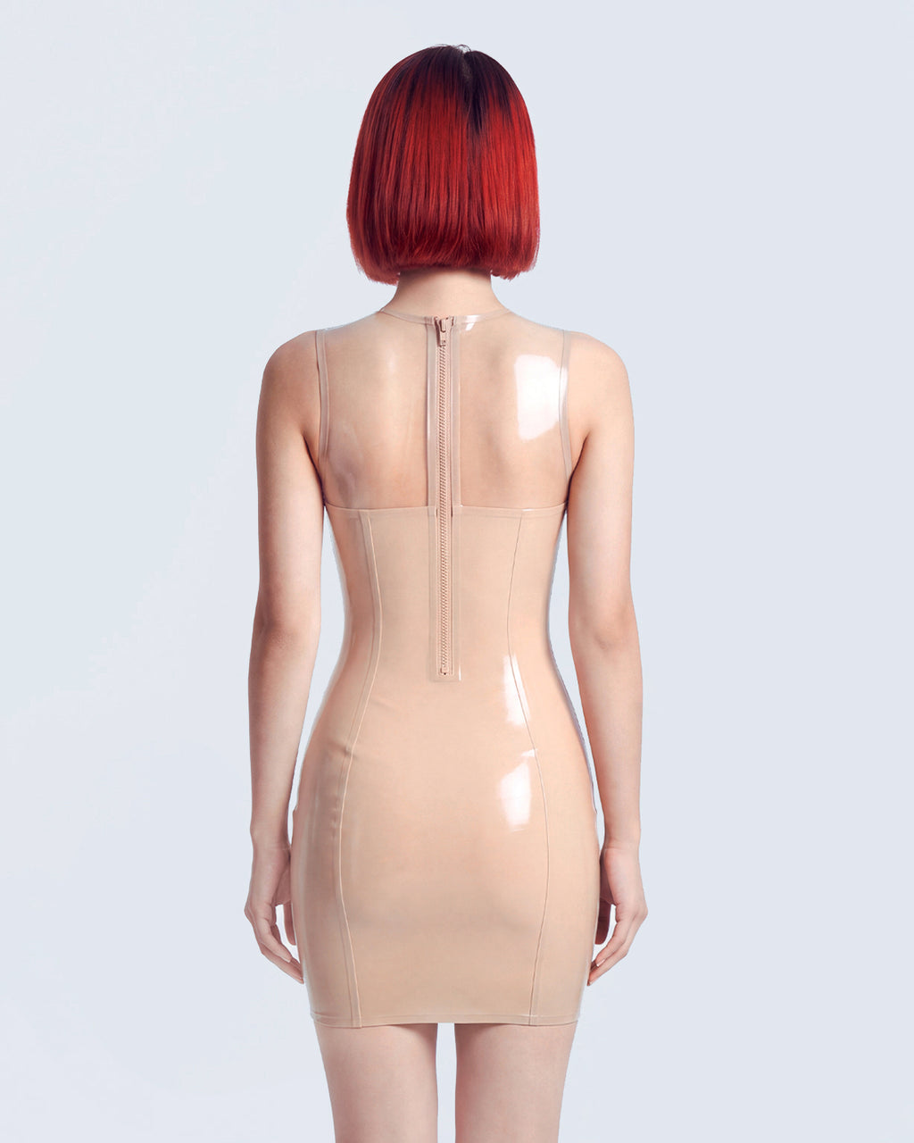 A model shown from the back wearing a high-shine nude latex dress by Ribcage, featuring a scoop neckline, sleeveless sheer transparent shoulders, opaque body panels, and a compressive second-skin pencil silhouette.