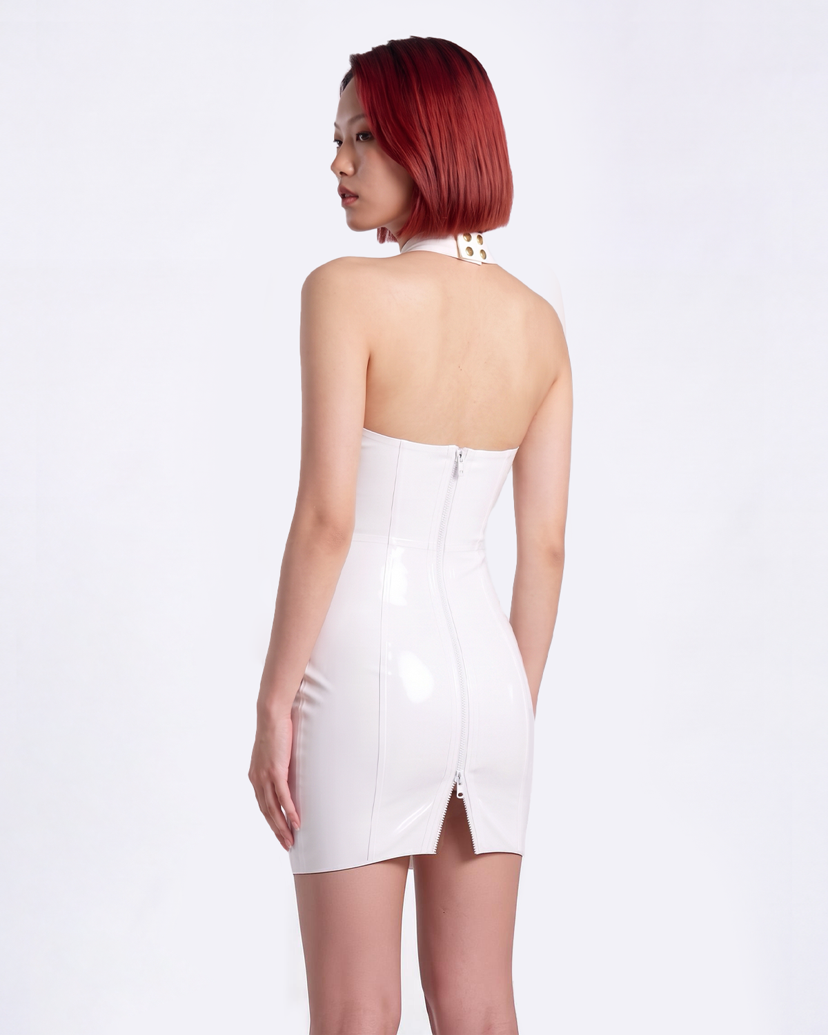 A model shown from the back wearing a high-shine white latex halter-neck dress by Ribcage, featuring an open back design, a high-neck collar, a prominent central zipper, and a compressive second-skin pencil silhouette.