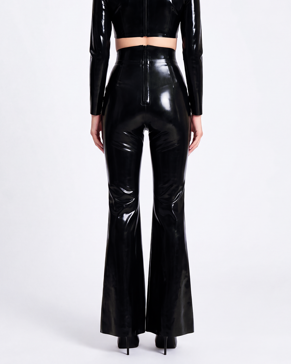 A model shown from the back wearing a pair of glossy metallic black latex high-waisted flared trousers by Ribcage, featuring a sharp architectural drape and a compressive second-skin fit that accentuates the silhouette.