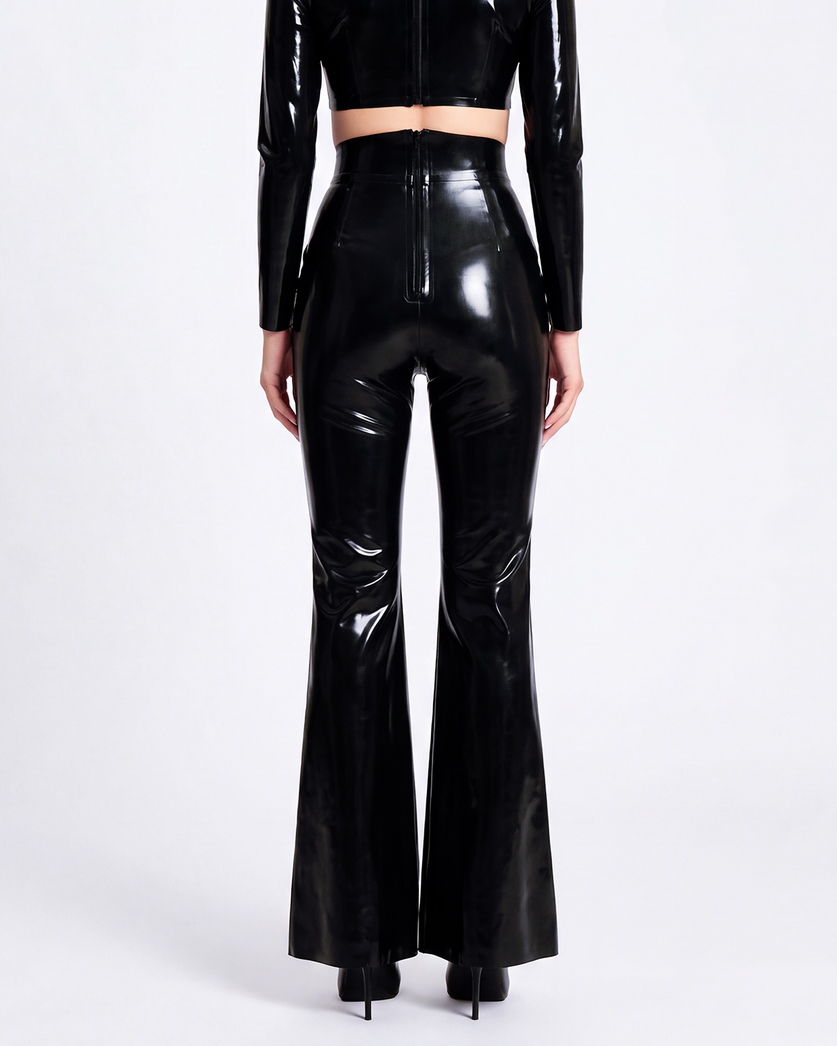 A model shown from the back wearing a pair of glossy metallic black latex high-waisted flared trousers by Ribcage, featuring a sharp architectural drape and a compressive second-skin fit that accentuates the silhouette.